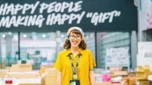 Lush Mitarbeitende Person in einem gelben T-Shirt lächelt und steht vor einem Schild auf dem steht: Happy People making happy gifts 