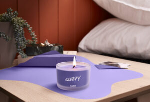 Die Sleepy Kerze steht auf einem Nachtkasterl neben einem Bett mit weißem Überzug. Die Kerze ist angezunden.