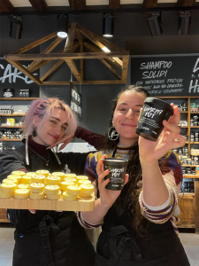 Due ragazze dello staff di Lush Venezia mostrano due prodotti Charity Pot, le mini coin solide e la confezione di crema per il corpo.