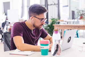 A person wearing a purple shirt, glasses and earphones looks attentively at their laptop in a bright office