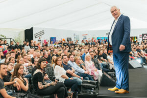 Mark Constantine stands on a stage in a white marquee in front of a large audience of Lush colleagues