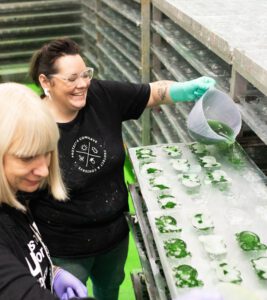 Two Lush manufacturing stand in the factory. The person in the foreground has light blonde hair and the person in the background has brown hair and is pouring liquid product into moulds to set.