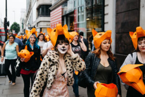 A group of fox hunting protesters walk down a street wearing orange fox masks on top of their heads. Two people are in the foreground and the person on the left has their face painted like a fox 