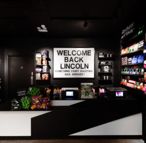 A white lightbox reads 'Welcome Back Lincoln' 'Something fairy exciting has arrived', in the middle of a darkly painted shop