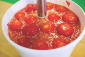 A white bucket filled with fresh tomatoes being mixed with an immersion blender