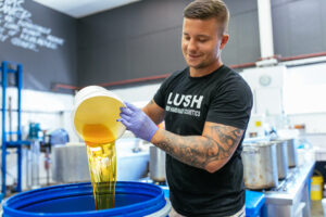 A Lush compounder pouring ingredients into a large blue barrel inside a Lush production room.