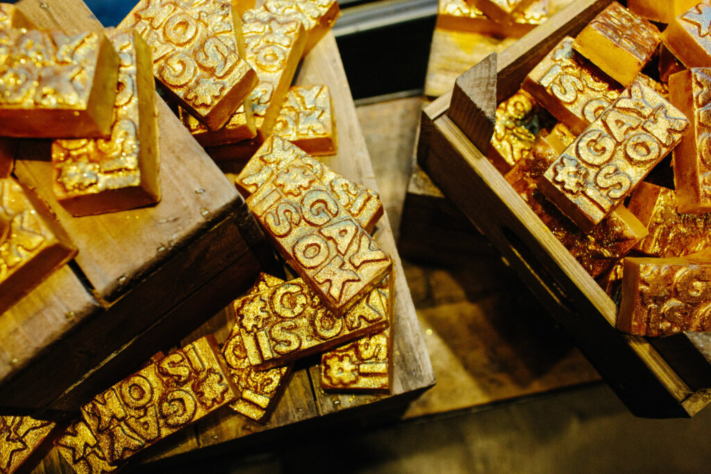 Multiple bars of gold #GayIsOk Soap are displayed randomly on wooden blocks and in wooden crates.