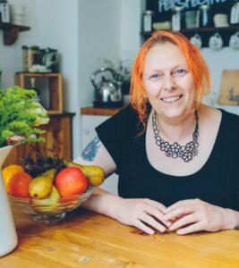Lush ethics director, Hilary sits at the Lush Spa kitchen table. Hilary has bright orange hair and is wearing black clothing while smiling