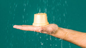 Cream coloured, top hat shaped Ro's Argan naked body conditioner is held under running water by an outstretched hand, against a teal background.