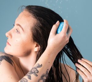 Against a light blue background and under a shower, a person with light skin tone, long dark hair and tattoos runs a blue coloured Big pressed conditioner bar through the lengths of their hair.