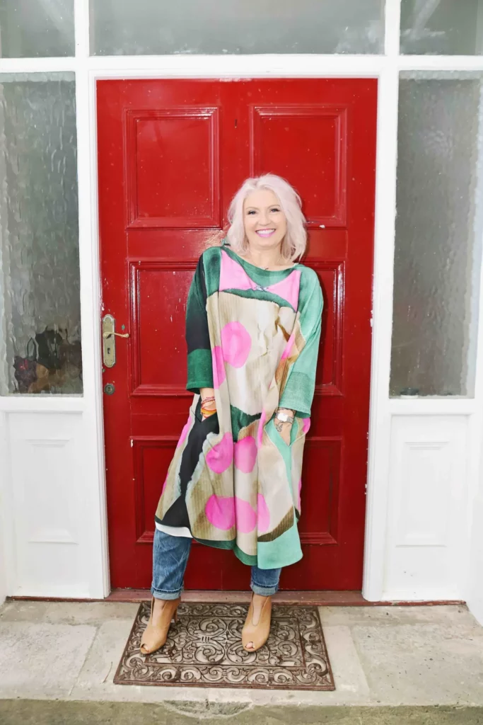 Photo of Rowena Bird smiling, wearing a colourful outfit, standing against a red door