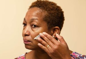 A person with medium skin tone and short afro curly hair looks thoughtful as they rub Self-Preserving Skin’s Shangri La moisturiser onto their cheek, using circular motions.