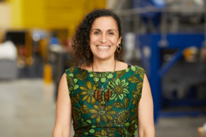 A Lush Colleague stands smiling in the Green Hub. They are wearing a green patterned dress, a handmade beaded necklace, and dark curly hair.