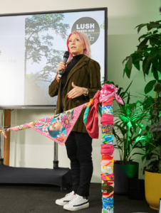 Lush Co-Founder Ro is stood on the stage at the opening of the Green Hub. She has pink hair and is wearing a gold patterned blazer, dark trousers and white trainers. She is surrounded by leafy plants.